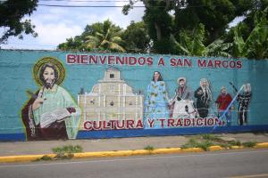 Bemalte Straßenmauer in San Marcos mit dem Schriftzug "Bienvenidos a San Marcos - Cultura y Tradición". Links ist Jesu Christu mit der Bibel zu sehen, daneben wichtige kulturelle Figuren aus dem Leben in San Marcos