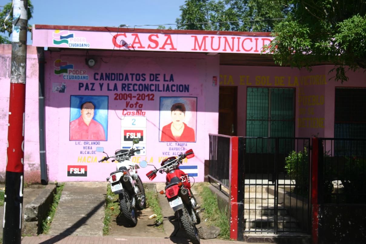 Zu sehen ist das Büro der Regierungspartei FSLN 2008 in San Marcos. Es ist einstöckig, rosa bemalt und trägt Wandbilder der damaligen Kandidaten für die anstehende Bürgermeisterwahl. Links sieht man Orlando Vega, der die Wahl gewann
