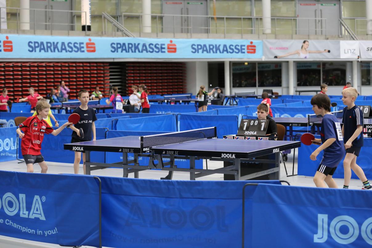 Junge Nachwuchs-Tischtennisspieler spielen in Zweier-Teams gegeneinander. Im Hintergrund sieht man weitere Kinder, die in einer Sporthalle gegeneinander antreten. 