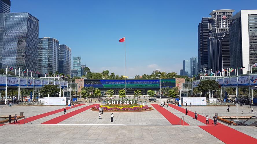 Der Haupteingang zur Hi-Tech Messe 2017 in Panyu. Davor ein weitläufiger Platz mit gepflegten Blumenbeet und der wehenden China-Flagge. Linker und rechter Bildrand werden von den markanten Wolkenkratzern der Stadt gesäumt