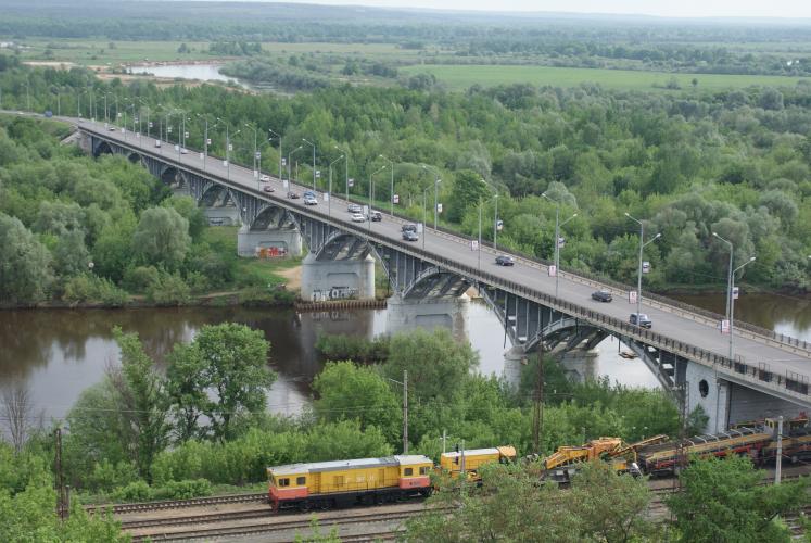 Zu sehen ist der Fluss Kljasma in einer grünen Ebene. Er wird von einer Autobrücke überspannt. Davor rollt gerade ein gelber Zug über die Schienen. 