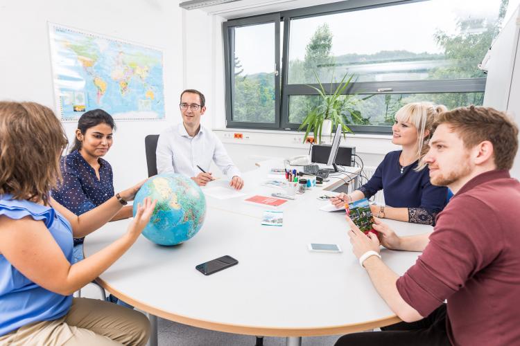 Studenten der EAH Jena sitzen um einen runden Tisch und unterhalten sich über internationale Angelegenheiten, symbolisiert durch einen Globus und eine Weltkarte, die an der Wand im Hintergrund des hellen Raumes zu sehen ist