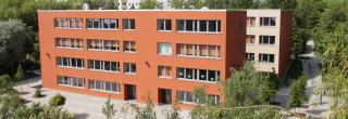 Das Schulgebäude der Grundschule Friedrich Schiller Jena ist in einem freundlichen orange-gelb-Ton gehalten. Die Schule ist von grünen Wiesen und Bäumen umrahmt. Im Hintergrund befindet sich ein Gebäudekomplex mit Wohnungen