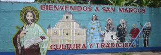 Bemalte Straßenmauer in San Marcos mit dem Schriftzug "Bienvenidos a San Marcos - Cultura y Tradición". Links ist Jesu Christu mit der Bibel zu sehen, daneben wichtige kulturelle Figuren aus dem Leben in San Marcos