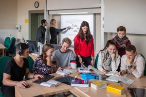 Schüler und Schülerinnen der Stoyschule sitzen und stehen um einen Tisch und sehen sich gemeinsam Unterrichtsmaterialien an. Im Hintergrund des modern ausgestatteten Klassenzimmers sieht man zwei weitere Schüler, die eine Grafik auf der interaktiven Tafel auswerten.