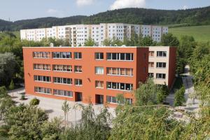 Das Schulgebäude der Grundschule Friedrich Schiller Jena ist in einem freundlichen orange-gelb-Ton gehalten. Die Schule ist von grünen Wiesen und Bäumen umrahmt. Im Hintergrund befindet sich ein Gebäudekomplex mit Wohnungen