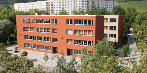 Das Schulgebäude der Grundschule Friedrich Schiller Jena ist in einem freundlichen orange-gelb-Ton gehalten. Die Schule ist von grünen Wiesen und Bäumen umrahmt. Im Hintergrund befindet sich ein Gebäudekomplex mit Wohnungen