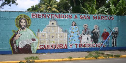 Bemalte Straßenmauer in San Marcos mit dem Schriftzug "Bienvenidos a San Marcos - Cultura y Tradición". Links ist Jesu Christu mit der Bibel zu sehen, daneben wichtige kulturelle Figuren aus dem Leben in San Marcos